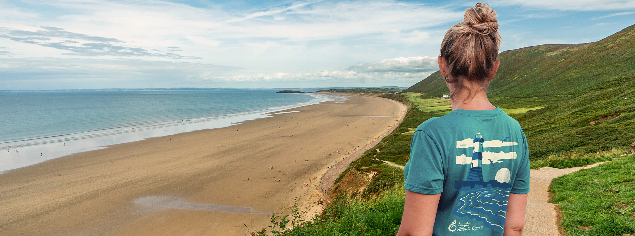 Woman wearing an official Wales Coast Path t-shirt looking out at the sea from the path.